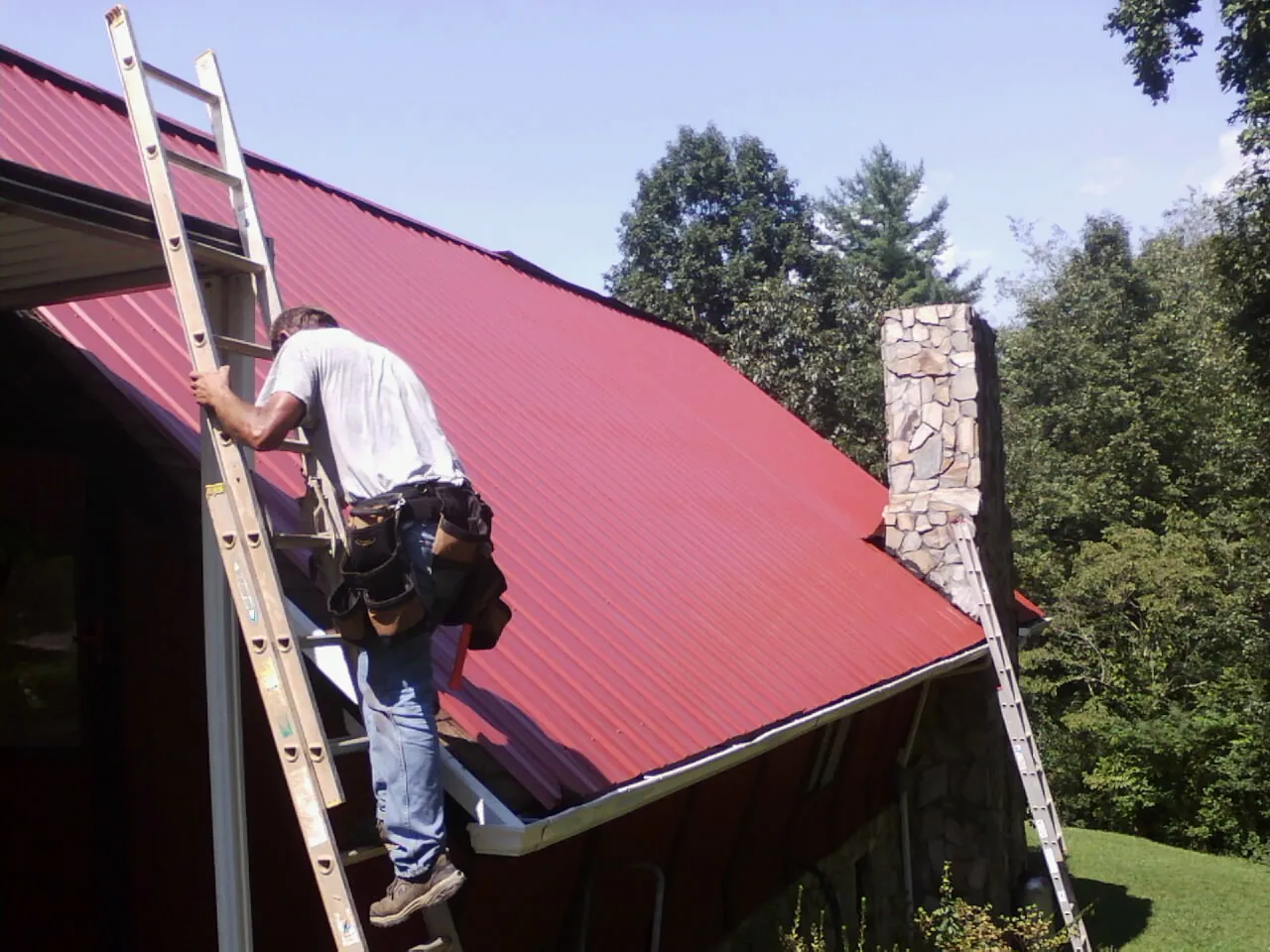 Roofer on a ladder beside a red metal roof and stone chimney — Soffit & Fascia Repair by Roofing Brothers in San Leandro